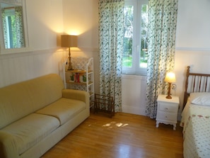 Ein gemütliches Zimmer mit Holzboden, einem beigefarbenen Sofa, einem kleinen weißen Nachttisch mit Lampe, einem Holzkopfende und einem Fenster mit floralen Vorhängen.