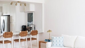 Uma sala de estar moderna com um sofá branco, uma mesa de centro e uma área de jantar com cadeiras, além de uma cozinha ao fundo.