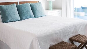 Quarto com cama, duas cadeiras de vime, mesa de cabeceira com abajur, ventoinha no teto e vista para o exterior.