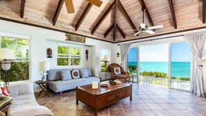 A spacious living room with a wooden ceiling, a large window overlooking the ocean, a sofa, a coffee table, and a chair.