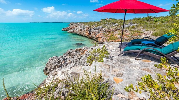 A beachside lounge with a red umbrella, stone flooring, and turquoise sea views.