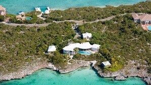 An aerial view of a tropical island with multiple buildings, including a large house with a swimming pool, surrounded by lush greenery and clear turquoise waters.