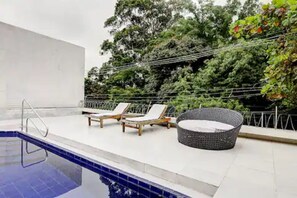 A rooftop pool area with lounge chairs and a wicker chair, surrounded by greenery and a white wall. - Casa Inteira (São Paulo)
