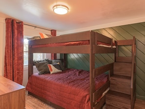 A bunk bed with a red quilt, a wooden desk, and a window with curtains.