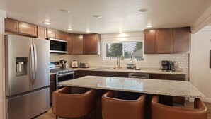 A modern kitchen with stainless steel appliances, wooden cabinets, a granite countertop, and three bar stools. - Big Yard Hot Tub Fire Pit Trail Pictured Rocks (Munising)