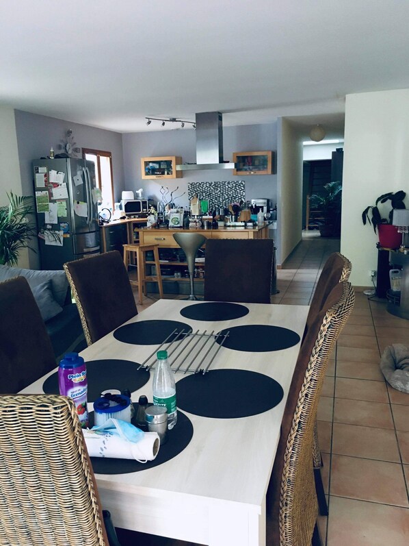 A dining area with a table set for four, surrounded by wicker chairs. A refrigerator and kitchen area are visible in the background. - Chambre Dans Maison Face à la Rivière Dain (Varambon)