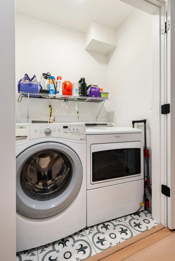 A compact laundry room with a front-loading washing machine, a drying machine, and a shelf above holding various cleaning supplies.