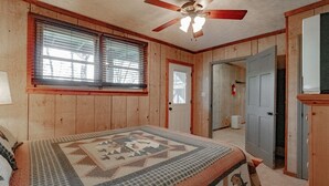 Une chambre à coucher comprenant un lit, un ventilateur de plafond, une fenêtre avec des stores et une porte donnant sur une autre pièce.