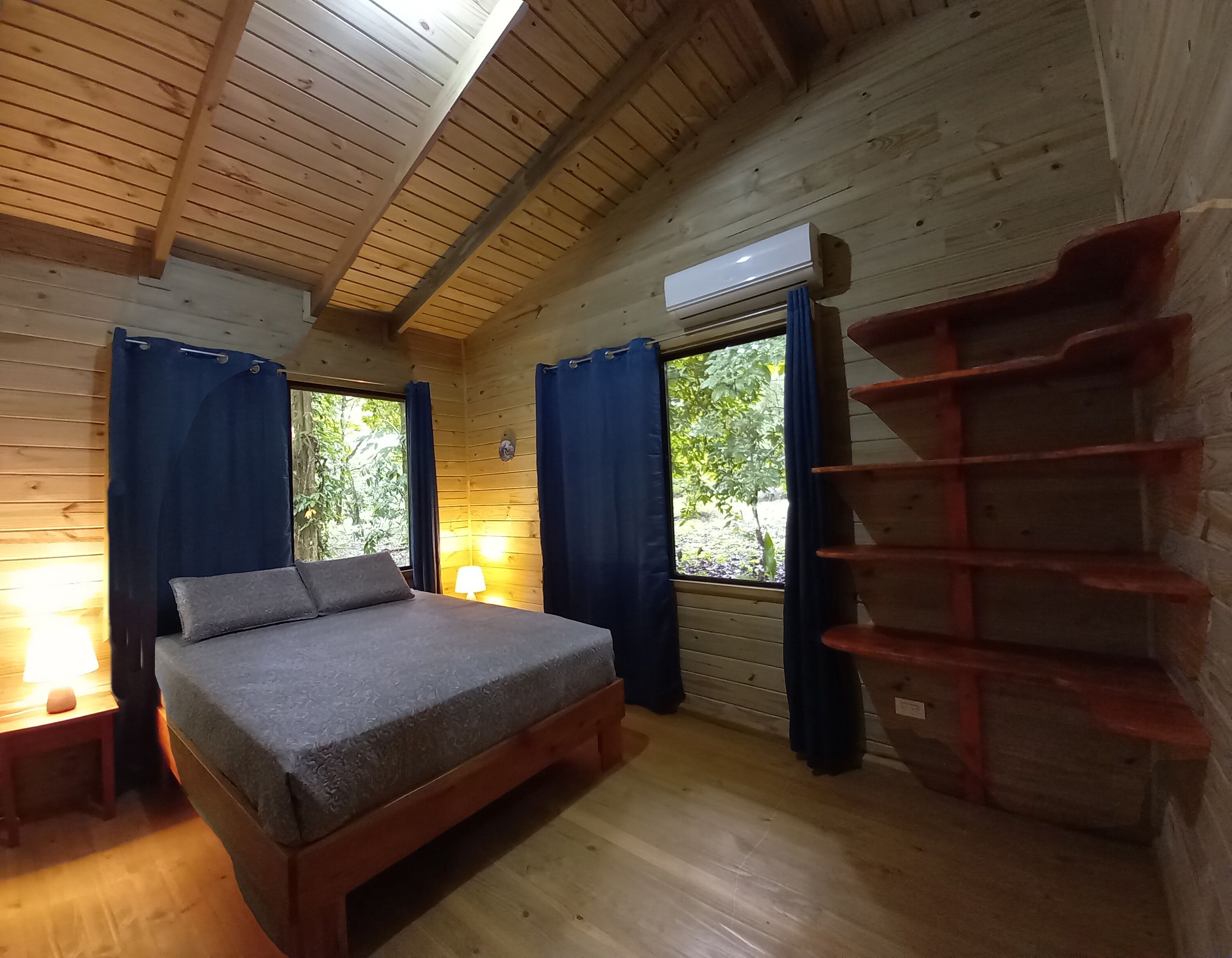 A wooden cabin room with a bed, two windows with curtains, a nightstand, and a wall-mounted shelf.