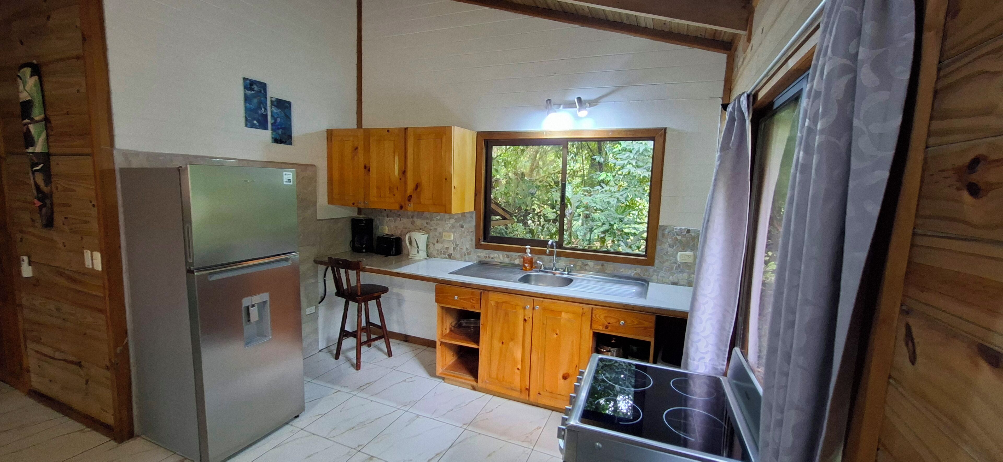 A compact kitchen with wooden cabinets, a refrigerator, a stove, and a window providing a view of greenery.
