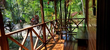 Una terraza de madera con barandilla da a un jardín exuberante con diversas plantas y un edificio pequeño al fondo.