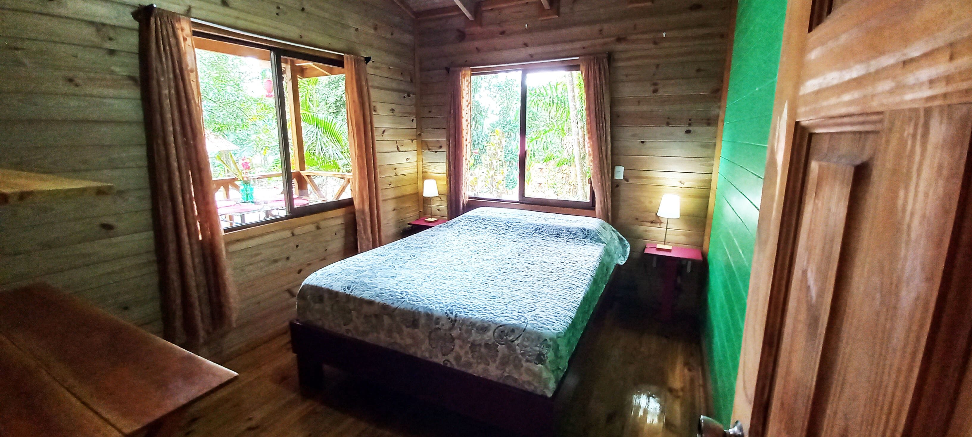A wooden cabin room with a bed, two windows, and a nightstand with a lamp.