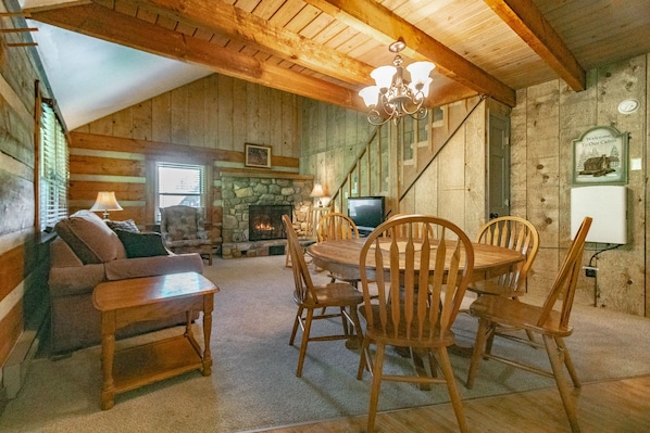 A cozy cabin interior with a stone fireplace, wooden dining table, and a television.
