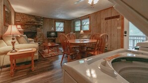 A cozy living room with a stone fireplace, wooden walls, a dining table with chairs, a sofa, a TV, and a hot tub.