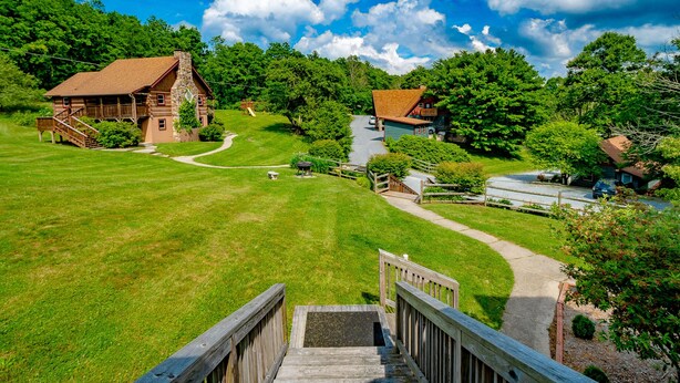 A rustic lodge with a stone chimney, a wooden deck, and a winding driveway surrounded by lush greenery.