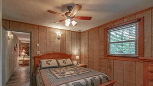 Un dormitorio con cama de madera, ventilador de techo, ventana con persianas y un pasillo que conduce a otra habitación.