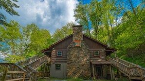 A log cabin with a stone chimney, wooden stairs, and a gravel driveway surrounded by trees.