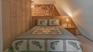 A bedroom with a sloped ceiling, wooden walls, a stone accent wall, a bed with a patterned bedspread, two pillows, and a bedside lamp. - Cozy Cabin #38 - Sleeps 6 - Full kitchen + fireplace - between Boone & Blowing Rock NC (Blowing Rock)