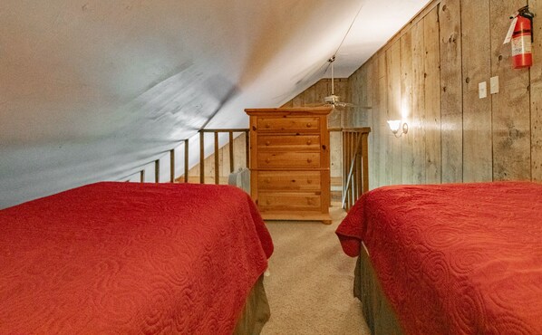 A small attic bedroom with two beds, wooden walls, a wooden dresser, a staircase, and a fire extinguisher.