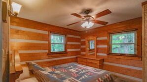 A bedroom with wooden walls, a bed with a quilt, a ceiling fan, and two windows with blinds.