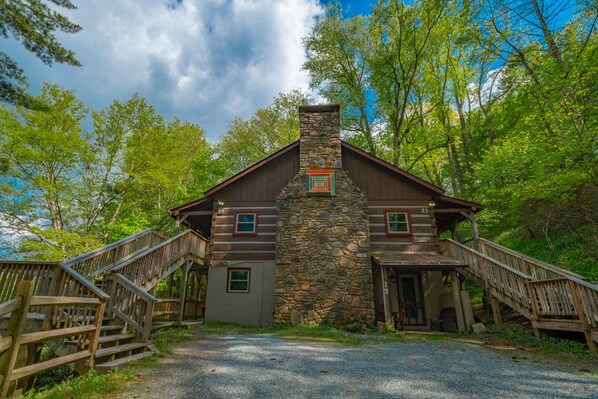 Une cabane en rondins avec une cheminée en pierre, un escalier en bois et une allée de gravier bordée d’arbres.