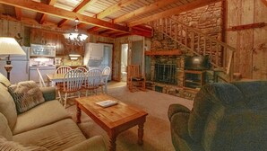 Uma sala de estar acolhedora com lareira, vigas de madeira, um espaço de jantar com cadeiras e uma cozinha ao fundo.