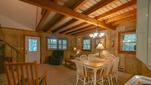 Interior acolhedor de uma cabana, com uma mesa de jantar de madeira, cadeiras brancas, lareira e uma janela grande com persianas.