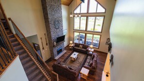 A spacious living room with a stone fireplace, a large window overlooking a deck, and a wooden staircase.