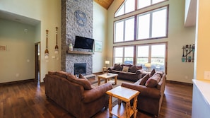 A spacious living room with a stone fireplace, large windows, and wooden flooring.