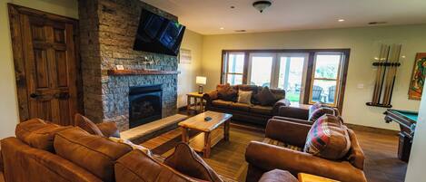 A cozy living room with a stone fireplace, leather sofas, a wooden coffee table, and a billiards rack.