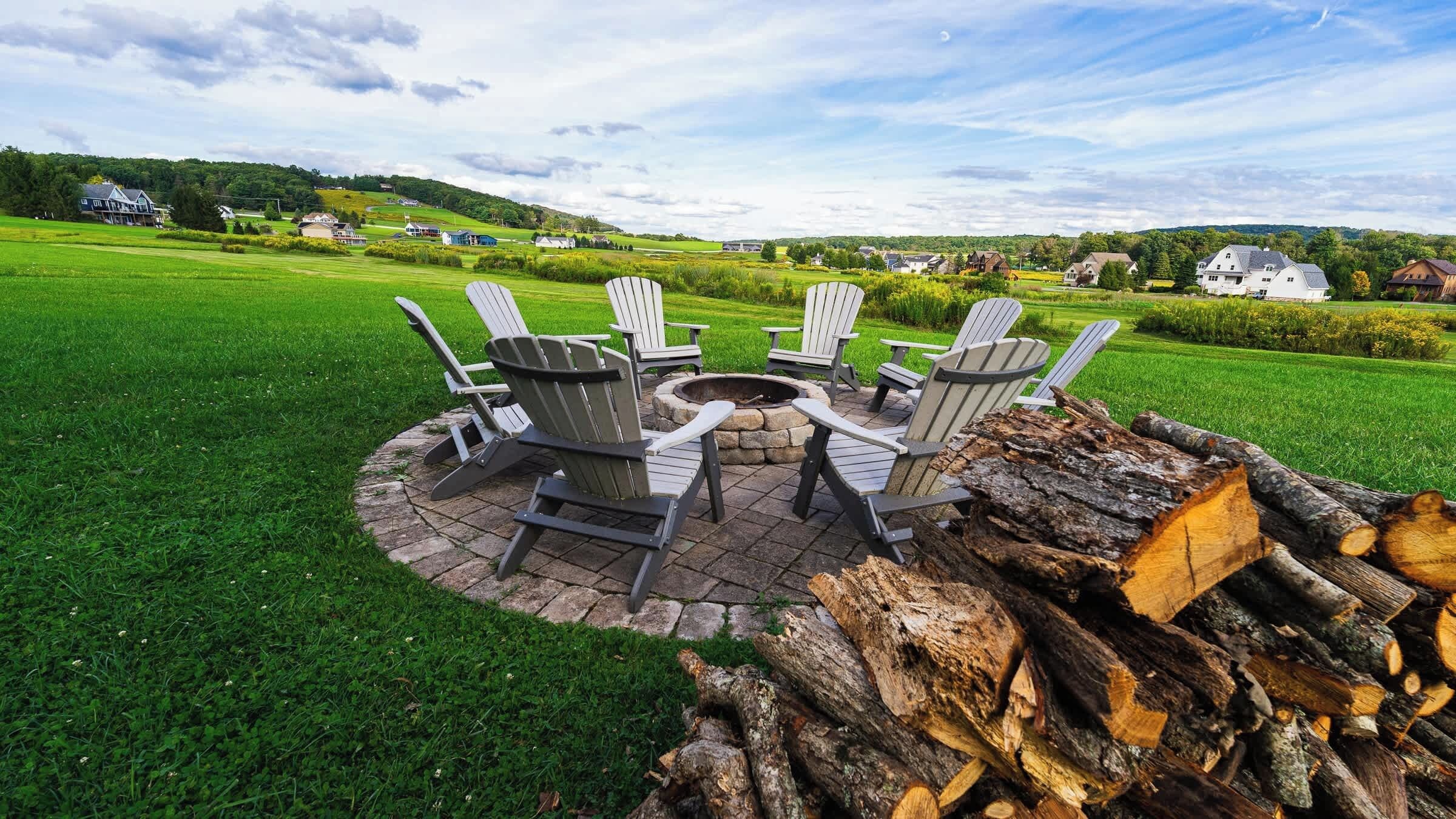 A circular outdoor seating area with Adirondack chairs around a fire pit, set on a stone patio with a backdrop of a green field and houses.
