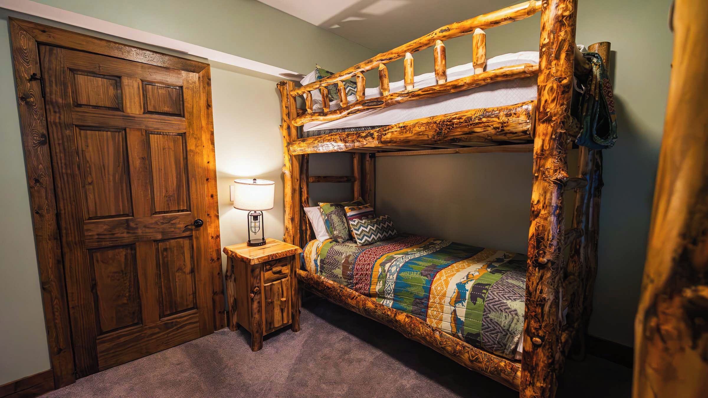 Una cama litera con estructura de madera, una mesita de noche pequeña con lámpara, y una puerta con diseño de paneles de madera.