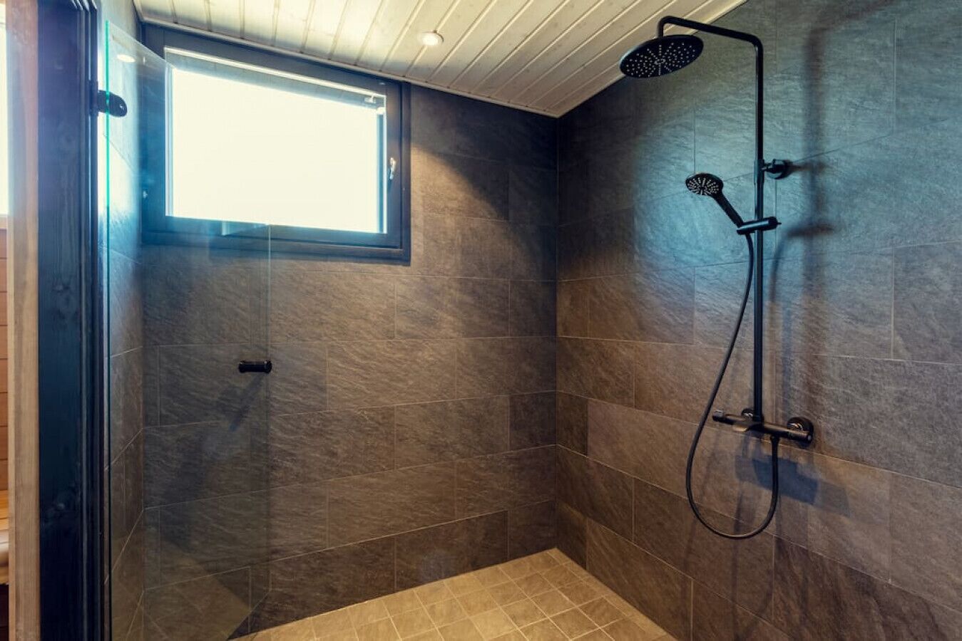Eine begehbare Dusche mit dunklen Fliesen, einem Fenster und einer Deckenleuchte.