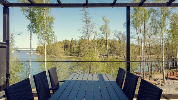 A covered outdoor seating area with a table and chairs, overlooking a lake and forested area with a bridge in the distance. - Villa Eslahti - Seafront/Jacuzzi (Porvoo)