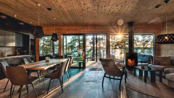 Un intérieur moderne avec un plafond en bois, un coin repas, une cheminée et de grandes fenêtres donnant sur la forêt.