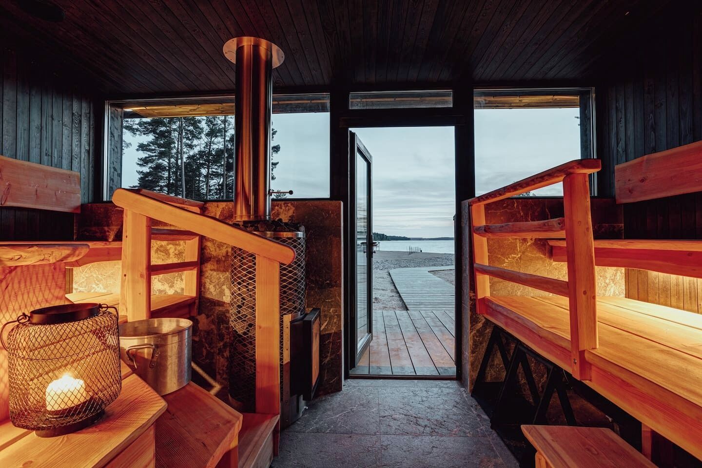 Interior acogedor de una cabaña con un estufa de leña central, bancos de madera y vista a una playa a través de un ventanal.