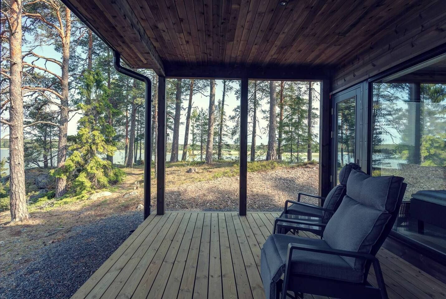 A covered deck with wooden flooring and two black chairs, overlooking a forested area with a lake.