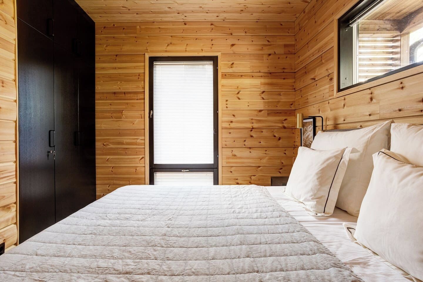 A bedroom with a wooden ceiling, a bed with white bedding, a window with blinds, and a dark wardrobe.