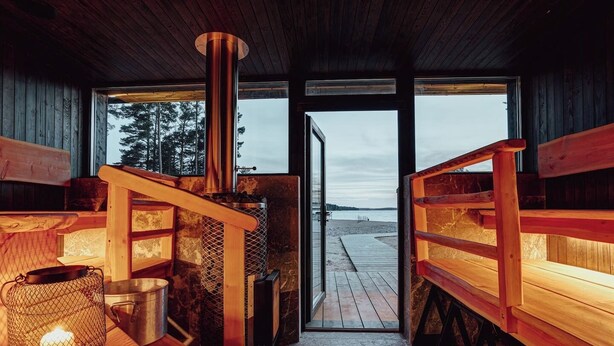 Interior acogedor de una cabaña con un estufa de leña central, bancos de madera y vista a una playa a través de un ventanal.