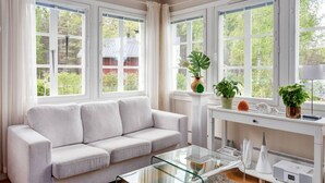 A modern living room with a light grey sofa, a glass coffee table, and a white console table with decorative items. Large windows provide a view of greenery outside.