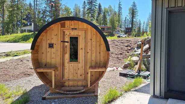 A wooden sauna with a door and a small window, surrounded by gravel and greenery, with a building and trees in the background. - Red Mtn Private Mountain Escape for Active Couples (Rossland)