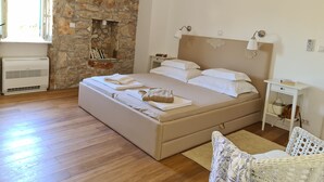 A bedroom with a stone wall, a bed with white linens, a bedside table with a lamp, and a small shelf with books.