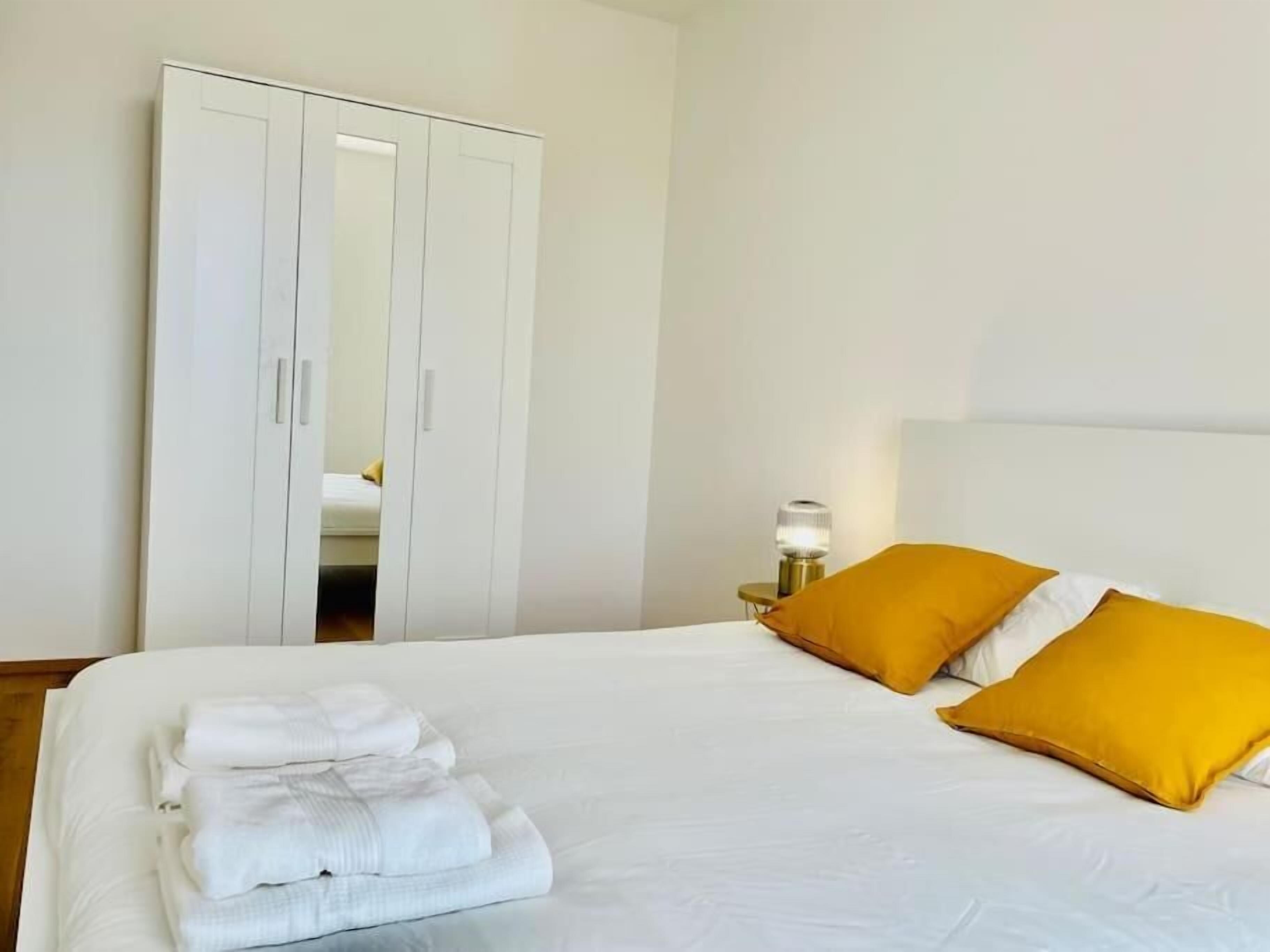 A neatly made bed with white linens and two mustard pillows, a bedside lamp, and a white wardrobe with a mirror.