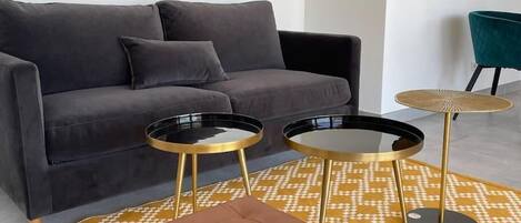 A living room with a grey sofa, two black and gold side tables, a brown ottoman, and a patterned rug.