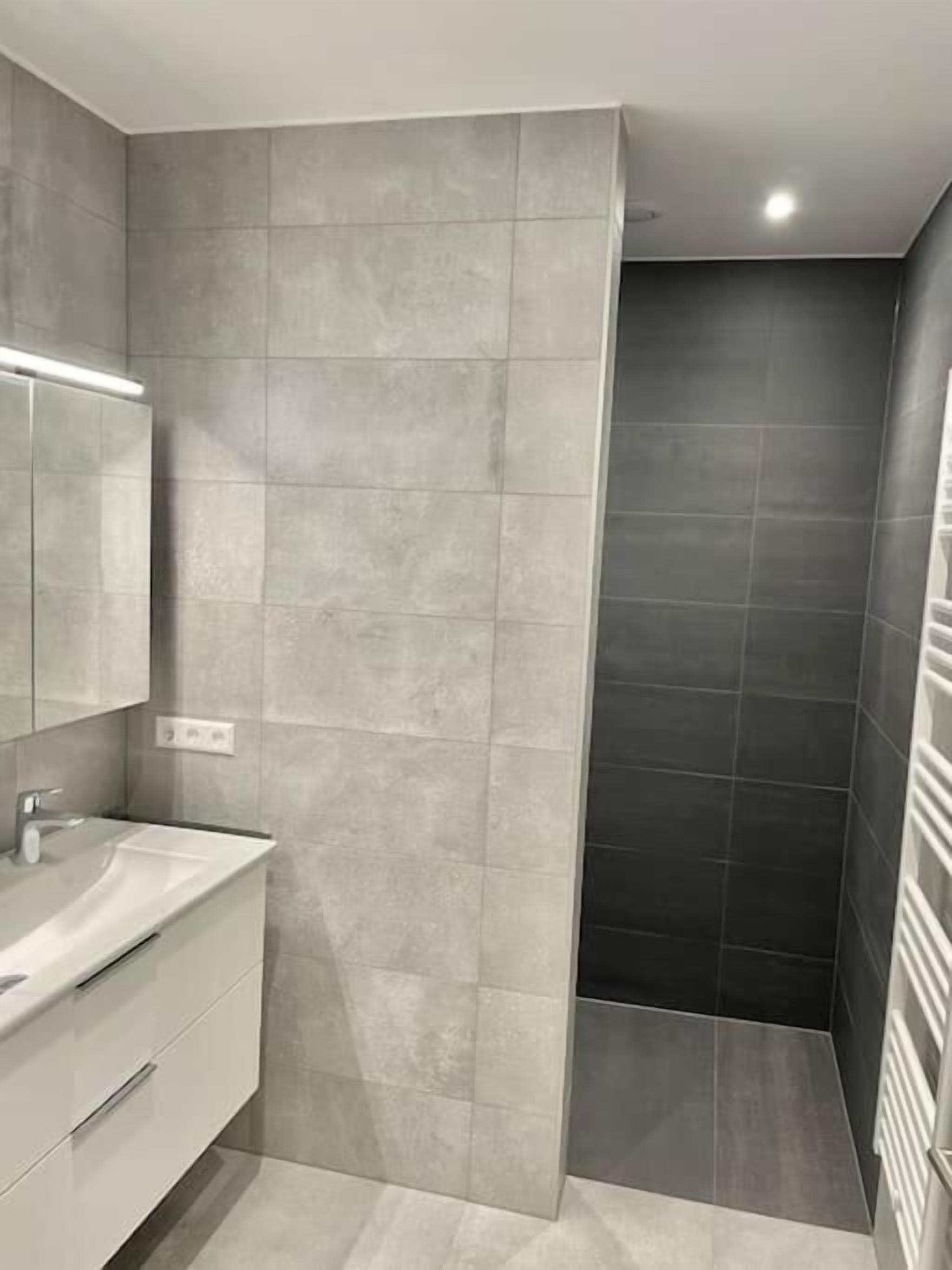 Un baño moderno con ducha de azulejos grises, lavamanos blanco con espejo y una lámpara en el techo.