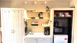 Cocina compacta con un refrigerador blanco, cafetera y microondas. Hay un mueble de madera con un lavamanos y una encimera con diversos utensilios de cocina.
