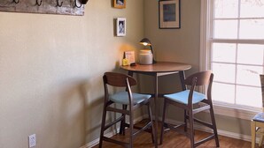 Una sala acogedora con piso de madera, un sofá gris, una mesa de comedor con dos sillas, una chimenea y una ventana con persianas.