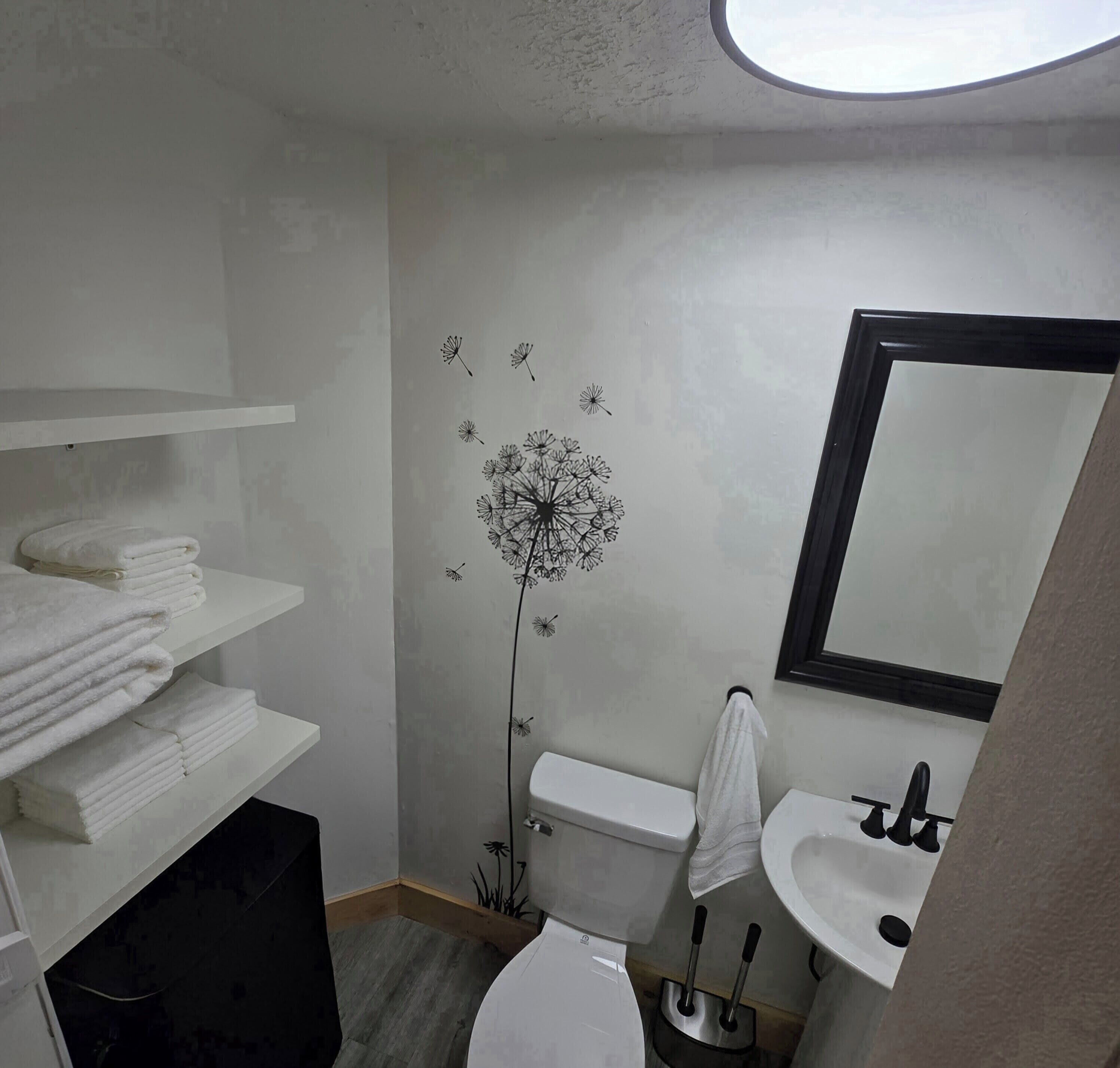 Een badkamer met toilet, wastafel en handdoekrek. Er zit een muurstickers van een paardenbloem op de muur. Planken zijn gevuld met witte handdoeken.
