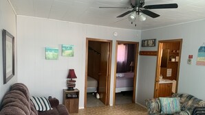 A living room with a brown sofa, a patterned sofa, and a wooden cabinet. There is a ceiling fan and a door leading to a bedroom.