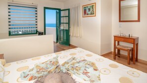Een slaapkamer met een bed, een houten nachtkastje, een raam met jaloezieën, een deur naar een balkon en een schilderij aan de muur.